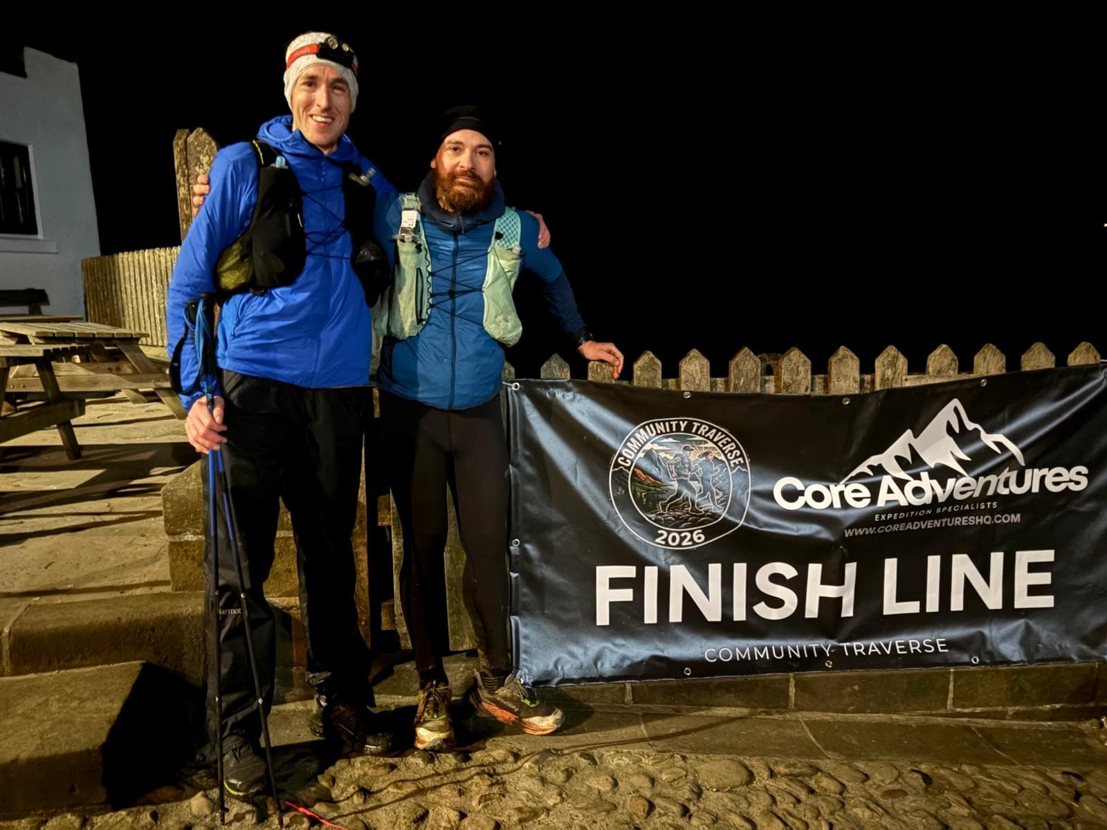 Two runners by the Community Traverse 2026 finish line banner after a night arrival